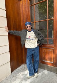 T-shirt beige surdimensionné avec lettrage bleu et rouge, veste bomber vert foncé, jean bleu décontracté, chaussons gris, bandana bleu, lunettes de soleil.