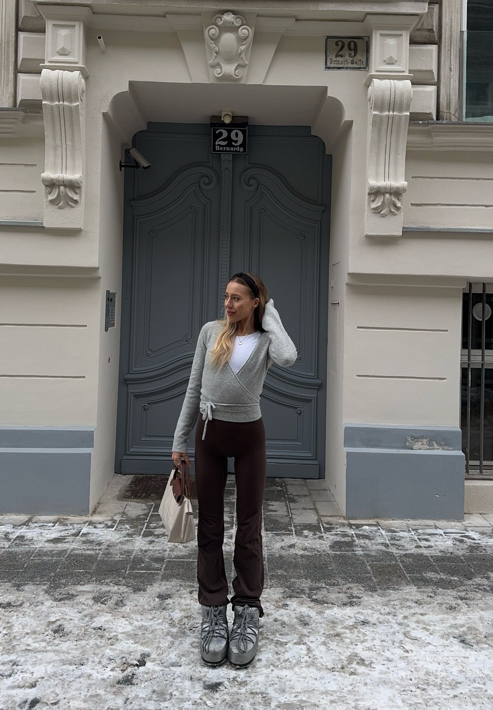Femme en pull gris enveloppant, pantalon marron et bottes argentées, debout dans une rue enneigée tenant un sac à main beige devant une porte grise ornée.