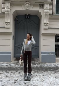 Femme en pull gris enveloppant, pantalon marron et bottes argentées, debout dans une rue enneigée tenant un sac à main beige devant une porte grise ornée.