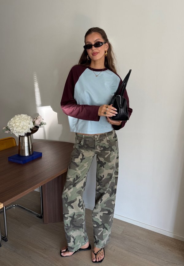 Femme portant des lunettes de soleil, une chemise bicolore à manches longues, un pantalon camouflage et des sandales, tenant un sac à main noir, debout à côté d'une table en bois avec des fleurs blanches.