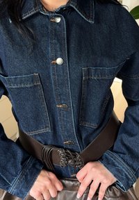 Jachetă din denim albastru închis cu închidere prin nasturi, două buzunare frontale și cusături contrastante; purtată cu o curea groasă maro.