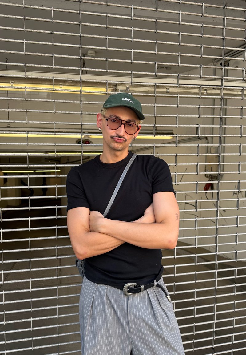 Jeune personne avec lunettes, t-shirt foncé, pantalon rayé et casquette verte, debout les bras croisés devant une grille métallique avec des lumières fluorescentes à l'intérieur.