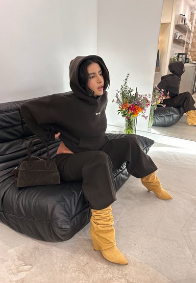 Hoodie marrón de gran tamaño, pantalones a juego, botas de tacón amarillas. Bolso negro al lado del modelo, que está sentado en un sofá de cuero negro con flores cerca.