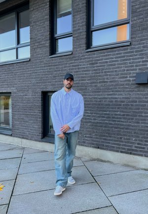 Camisa de botón de gran tamaño azul claro con un bolsillo, combinada con jeans de mezclilla holgados y descoloridos, y zapatillas grises, todo sobre un fondo de pared de ladrillos oscuros.