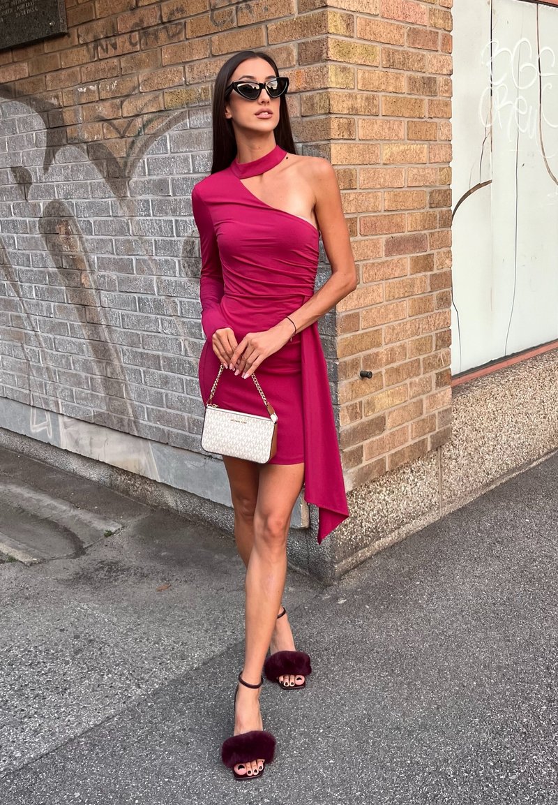 Mulher vestindo um vestido mini de uma só alça em magenta e óculos de sol pretos, segurando uma pequena bolsa branca contra uma parede de tijolos com grafite.