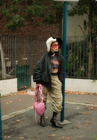 Person trägt schwarzen und weißen Bucket Hat, übergroße schwarze Jacke, Crop-Top mit Flammenmuster, khakifarbene Hose, schwarze Schnürstiefel und hält eine rosa herzförmige Tasche.