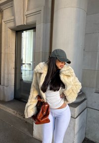 Crème jasje van imitatiebont met een brede kraag, gecombineerd met een wit overhemd en witte legging. Bruine leren handtas en donkere pet maken de look compleet.