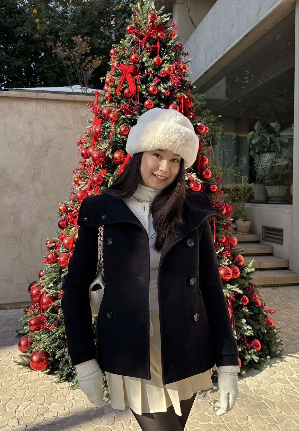 Cappotto nero con grandi bottoni, cappello di pelliccia bianca, maglione crema e gonna beige. In piedi davanti a un albero di Natale decorato con ornamenti rossi.
