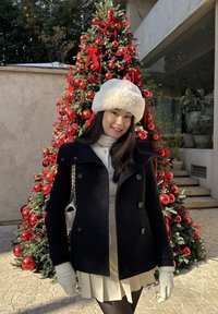 Cappotto nero con grandi bottoni, cappello di pelliccia bianca, maglione crema e gonna beige. In piedi davanti a un albero di Natale decorato con ornamenti rossi.