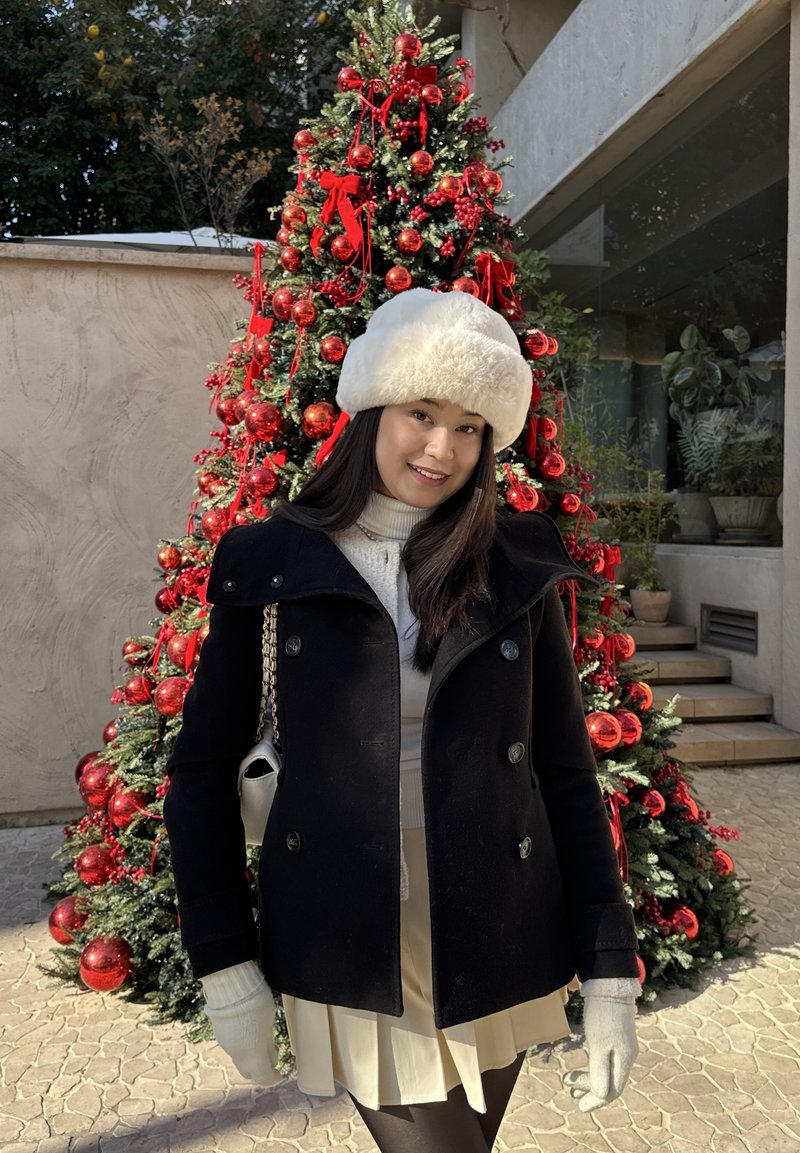 Manteau noir avec de gros boutons, chapeau en fourrure blanche, pull crème et jupe beige. Debout devant un sapin de Noël décoré avec des ornements rouges.