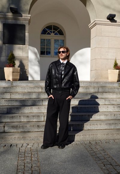 Hombre con chaqueta de cuero negra, pantalones negros anchos y gafas de sol, de pie sobre escalones de piedra frente a la entrada arqueada de un edificio.