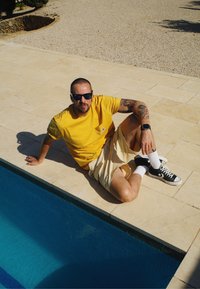 Camiseta de algodão amarela com bolso, calções bege, sapatos Converse pretos e meias brancas. Sentado ao lado de uma piscina azul em uma superfície de azulejos.