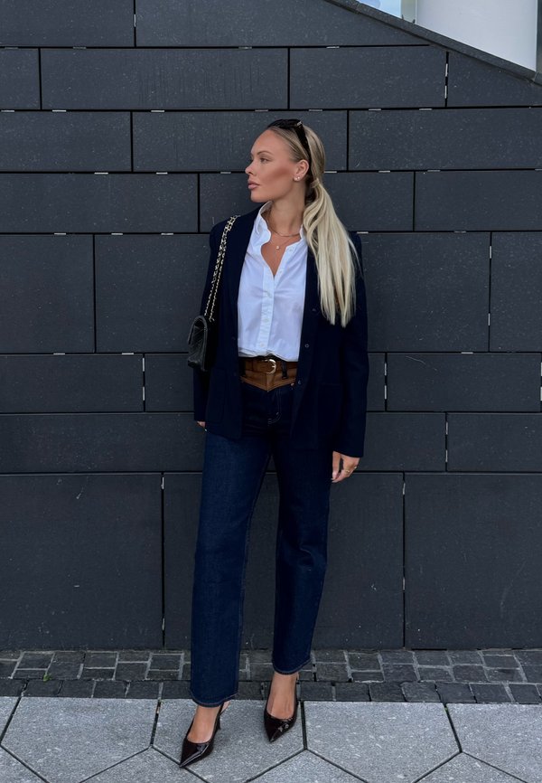 Donna con lunghi capelli biondi che indossa una camicia bianca, blazer blu navy, jeans scuri, cintura marrone, borsa nera e tacchi neri, in piedi contro un muro piastrellato scuro.