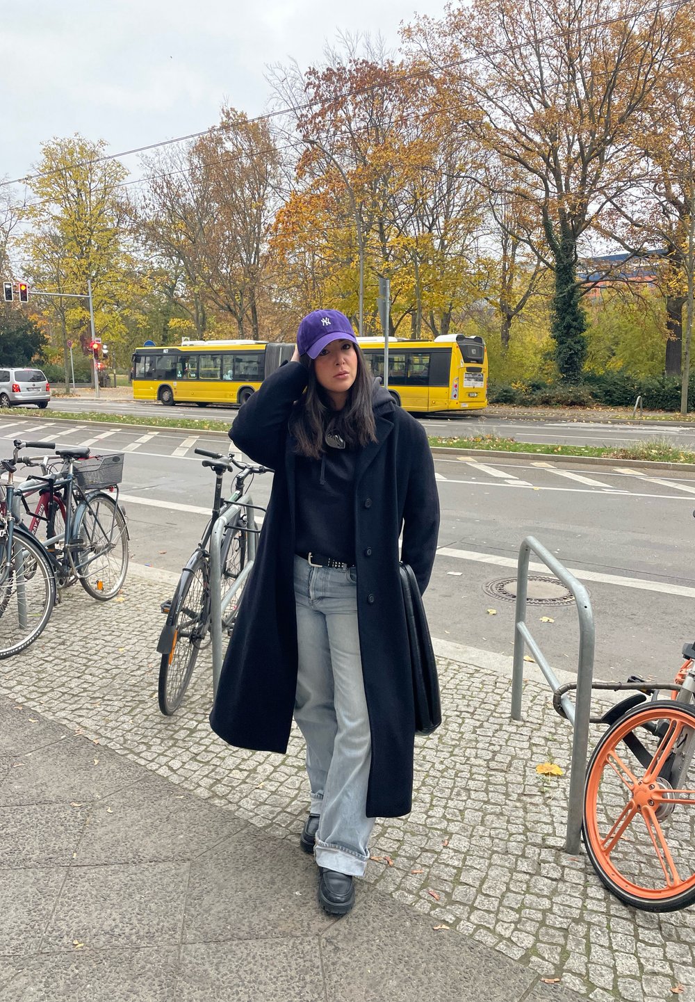 Μαύρο μακρύ παλτό, μοβ καπέλο με λογότυπο, μαύρη μπλούζα με κουκούλα, φθαρμένο ανοιχτό μπλε τζιν και μαύρες μπότες. Ποδήλατα και ένα λεωφορείο στο φόντο.