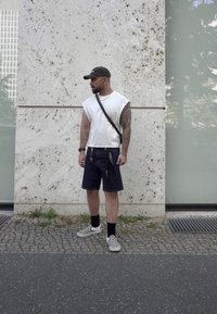 Weißes ärmelloses Shirt, marineblaue Shorts mit Verzierungen, schwarz-weiß kariertes Schuhwerk, schwarze Kappe, stehend an einer strukturierten Steinwand.