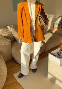 Oransje semsket blazer over en hvit topp, kombinert med vide hvite bukser og mørke slip-on sko. Holder en teksturert grå clutch.