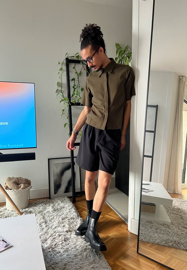 Homme aux cheveux bouclés attachés en chignon, portant des lunettes, une chemise à manches courtes olive, un short noir et des bottes noires, debout près d’un miroir dans un salon moderne.