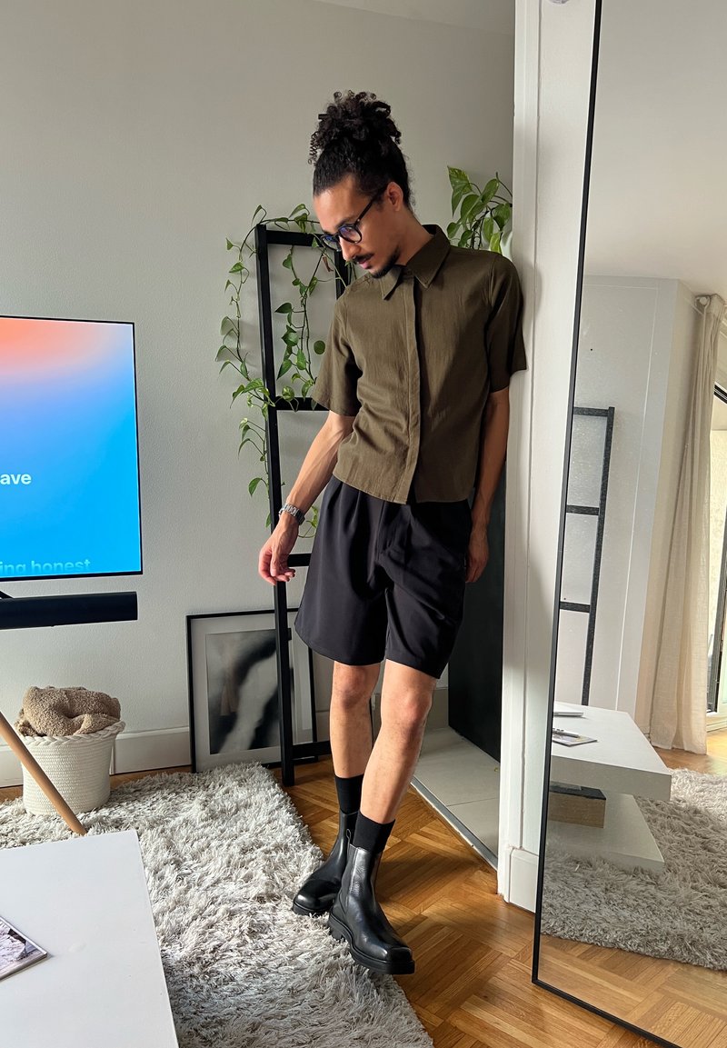 Homme aux cheveux bouclés attachés en chignon, portant des lunettes, une chemise à manches courtes olive, un short noir et des bottes noires, debout près d’un miroir dans un salon moderne.