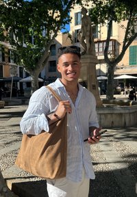 Saco de tote de camurça castanho, formato retangular, textura suave. Camisa de manga longa em azul claro e branco, de pé ao ar livre perto de uma estátua.
