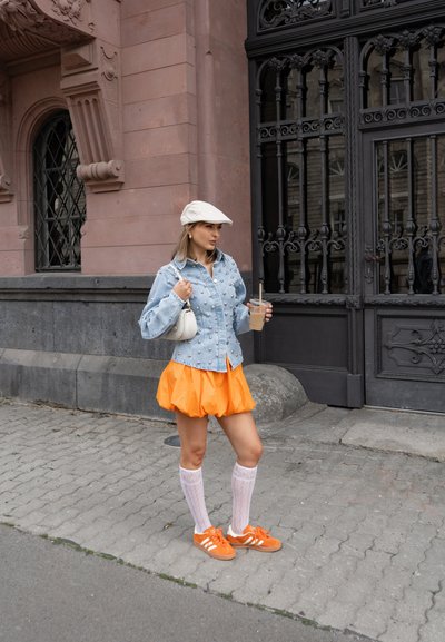 Mujer con falda naranja y zapatillas, chaqueta vaquera adornada con perlas, gorra blanca y calcetines hasta la rodilla sostiene una bebida junto a una puerta negra ornamentada.