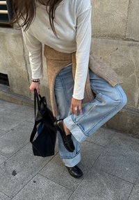 Czarne mokasyny, jasnoniebieskie dżinsy z podwiniętymi mankietami, biała koszula z długim rękawem oraz beżowy dziergany szal, trzymająca teksturowaną czarną torbę typu tote.