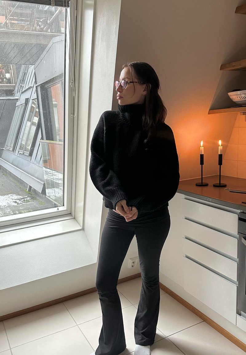 Maglione nero lavorato a maglia con collo alto, abbinato a pantaloni neri a zampa d'elefante. La stanza è caratterizzata da una grande finestra e due candele accese sul piano della cucina.