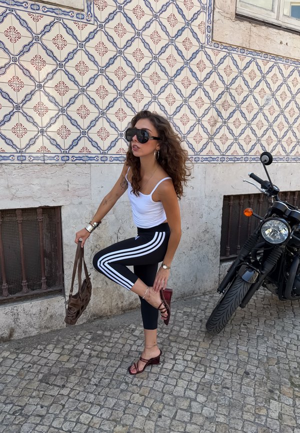 Donna con canotta bianca e leggings neri a righe si sistema la scarpa su una strada acciottolata vicino a un muro piastrellato a motivi e una moto parcheggiata.