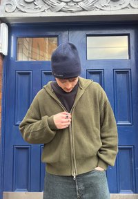 Ung mann i grønn strikket hettegenser og svart beanie som åpner jakken, står foran en blå dør med glasspaneler og utsmykket hvit trim.