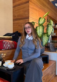 Jeune femme aux longs cheveux et lunettes assise à l'intérieur sur un banc en bois avec des tasses de café, une plante et des étagères avec des bouteilles en arrière-plan.