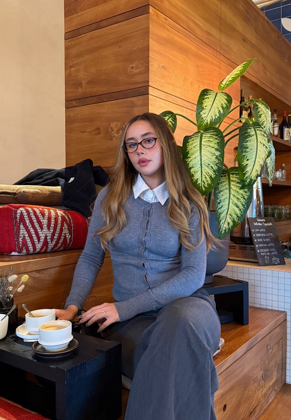 Jeune femme aux longs cheveux et lunettes assise à l'intérieur sur un banc en bois avec des tasses de café, une plante et des étagères avec des bouteilles en arrière-plan.