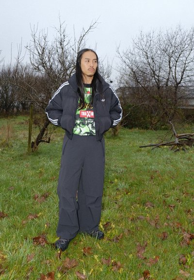 Chaqueta negra acolchada con rayas blancas, superpuesta sobre una camiseta gráfica con detalles verdes y rojos, combinada con pantalones grises anchos.