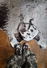 Kabelka z kravské kůže s zakřiveným tvarem, zdobená skvrnitým vzorem v bílé a hnědé barvě. Černobílé tenisky se podobným vzorem.