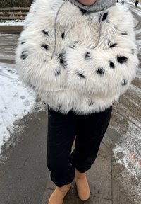 Personne portant une veste duveteuse blanche à taches noires, un pantalon noir, des bottes couleur fauve et une écharpe grise, debout sur un trottoir enneigé.