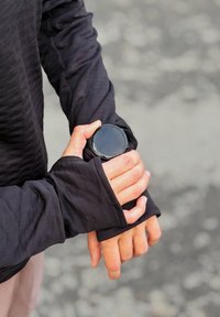 Czarna bluzka z długim rękawem wykonana z fakturowanej tkaniny, z otworami na kciuki. Na nadgarstku znajduje się czarny, okrągły zegarek z gładką szklaną tarczą.