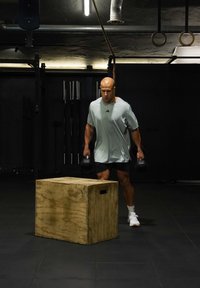 Homme en chemise claire et short tenant des haltères marchant vers une boîte plyométrique en bois dans une salle de sport faiblement éclairée avec des anneaux et des barres en arrière-plan.