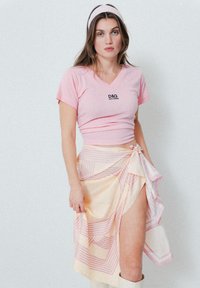 T-shirt rose à col en V avec logo D&G, associé à une jupe portefeuille à motifs jaune clair avec des rayures roses, et des bottes beiges montantes.