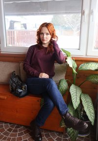 Jeune femme aux cheveux roux portant un pull bordeaux et un jean, assise sur un banc en bois près d'une fenêtre, à côté d'une plante verte en pot et d'un sac à main noir.