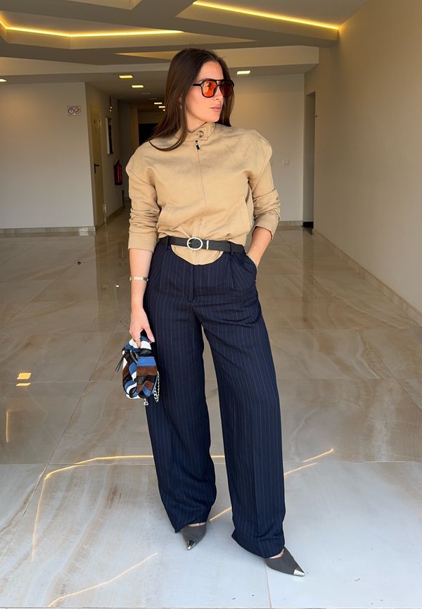 Femme portant une veste beige, ceinture noire, pantalon large bleu marine à rayures fines, chaussures grises pointues, lunettes de soleil orange, tenant un petit sac coloré à l'intérieur.