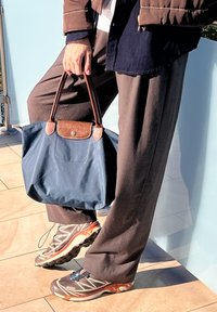 Nαυτικό μπλε τσάντα tote με καφέ δερμάτινες λεπτομέρε�ιες, συνδυασμένη με χαλαρά γκρι παντελόνια και γκρι αθλητικά παπούτσια με πορτοκαλί λεπτομέρειες.