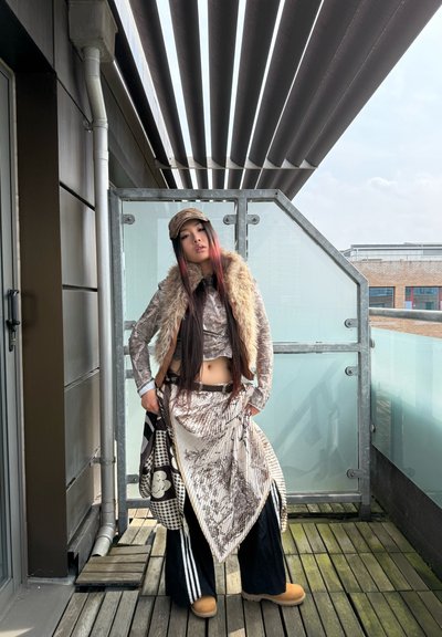 Chaqueta metálica con cuello de piel sintética, falda asimétrica con estampado, pantalones anchos negros, gorra y bolso de mano con detalles gráficos. Entorno al aire libre.