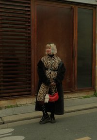 Leopardmønster kåpe over svart klær, tilbehørt med en rød veske med en fluffy charm, satt mot en trefon.