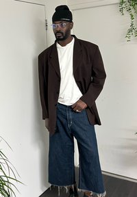 Veste blazer marron surdimensionnée, T-shirt blanc, jean denim à jambes larges avec ourlets effilochés, bonnet noir, lunettes rondes, debout contre un mur simple.