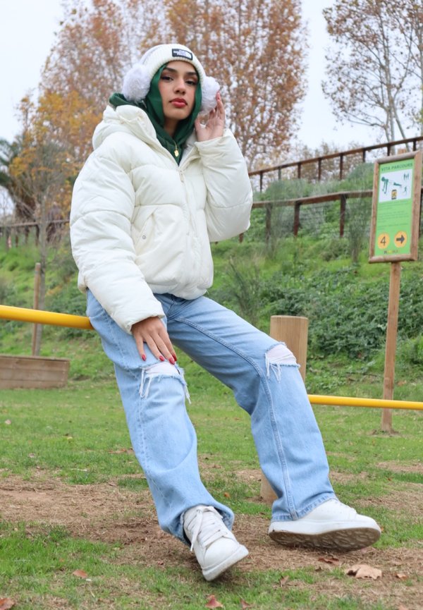 Chaqueta blanca hinchada, sudadera verde, vaqueros claros rasgados y zapatillas blancas. Modelo posando sobre hierba con una estructura de madera y señalización al fondo.