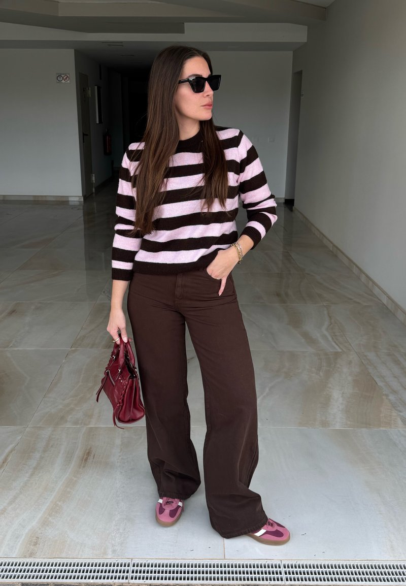 Femme portant des lunettes de soleil noires, un pull rayé rose et noir, un pantalon marron et des chaussures roses, tenant un sac à main rouge dans un couloir carrelé.