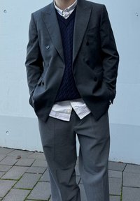 Γκρί σακάκι με διπλό κούμπωμα πάνω από γαλάζια πλεκτή αμάνικη μπλούζα, σε στρώσεις πάνω από λευκό και μπλε ριγέ πουκάμισο, σε συνδυασμό με γκρίζες παντελόνες.