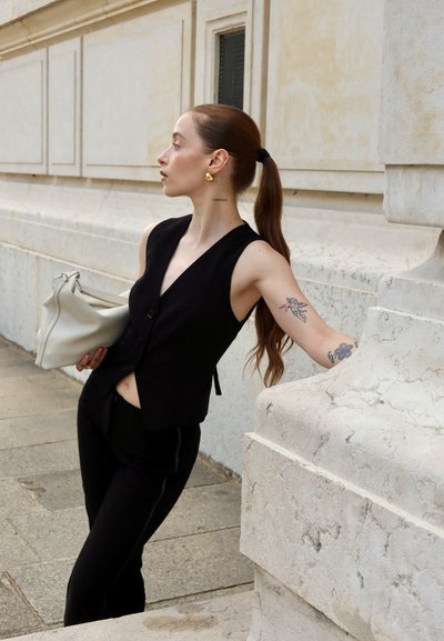 Chaleco sin mangas negro con botones, combinado con pantalones negros. Un bolso gris claro, pendientes de oro y tatuajes visibles en el brazo.