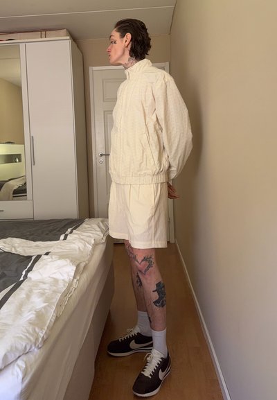 Persona con tatuajes en las piernas, de pie de lado, con una chaqueta beige y pantalones cortos en un dormitorio con una cama y un armario.