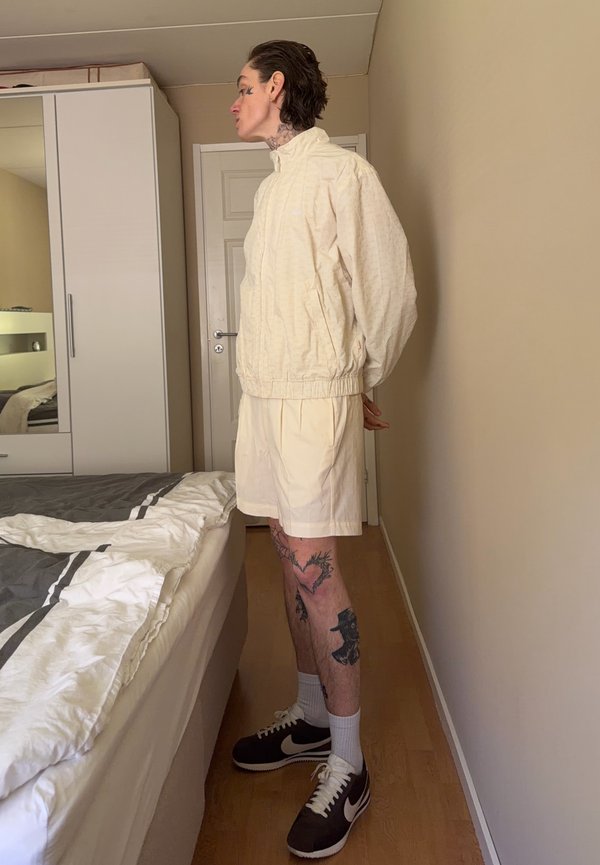 Personne avec des tatouages sur les jambes, debout de profil en veste beige et short dans une chambre avec un lit et une armoire.