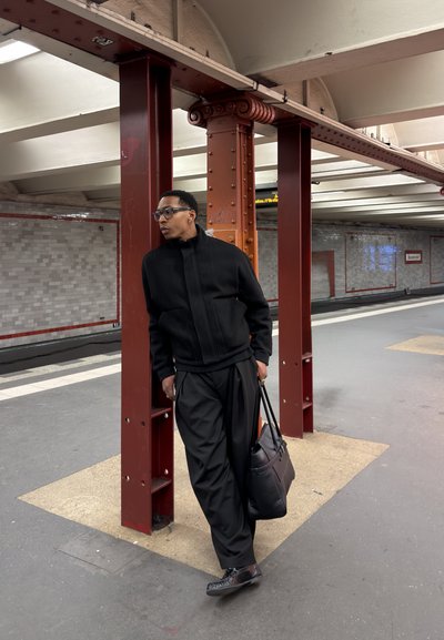 Hombre con chaqueta y pantalones negros apoyado en un pilar rojo en el andén del metro, sosteniendo una bolsa negra y mirando hacia un lado.