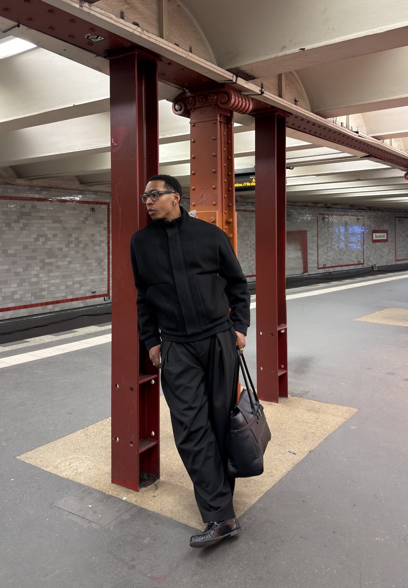 Homme en veste et pantalon noirs appuyé contre un pilier rouge sur le quai du métro, tenant un sac noir et regardant sur le côté.